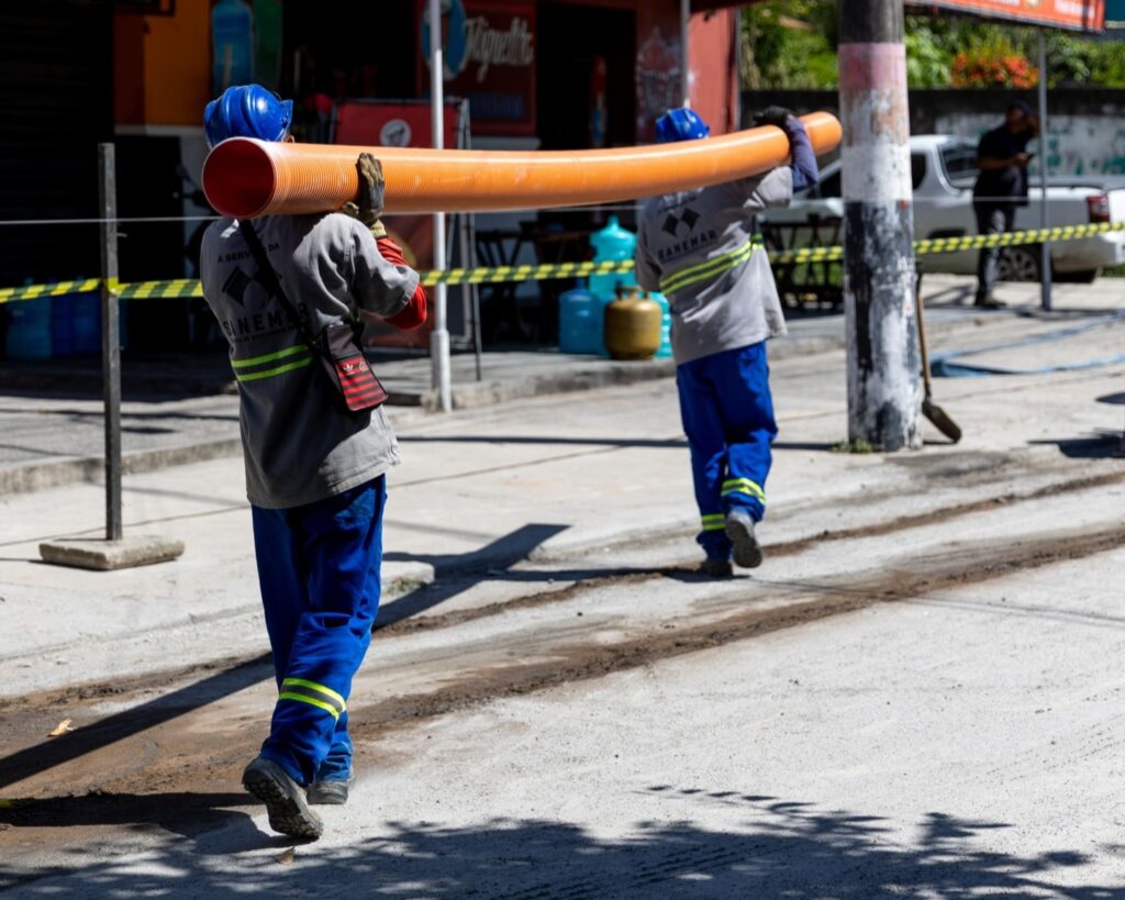 Obras de expansão da rede de esgoto em uma rua de Maricá, com maquinário e trabalhadores.