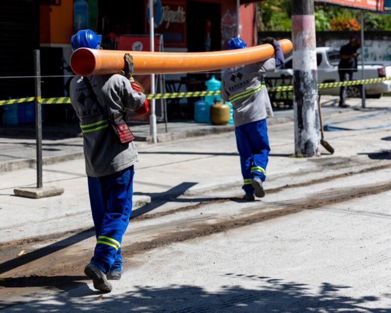 Obras de expansão da rede de esgoto em uma rua de Maricá, com maquinário e trabalhadores.