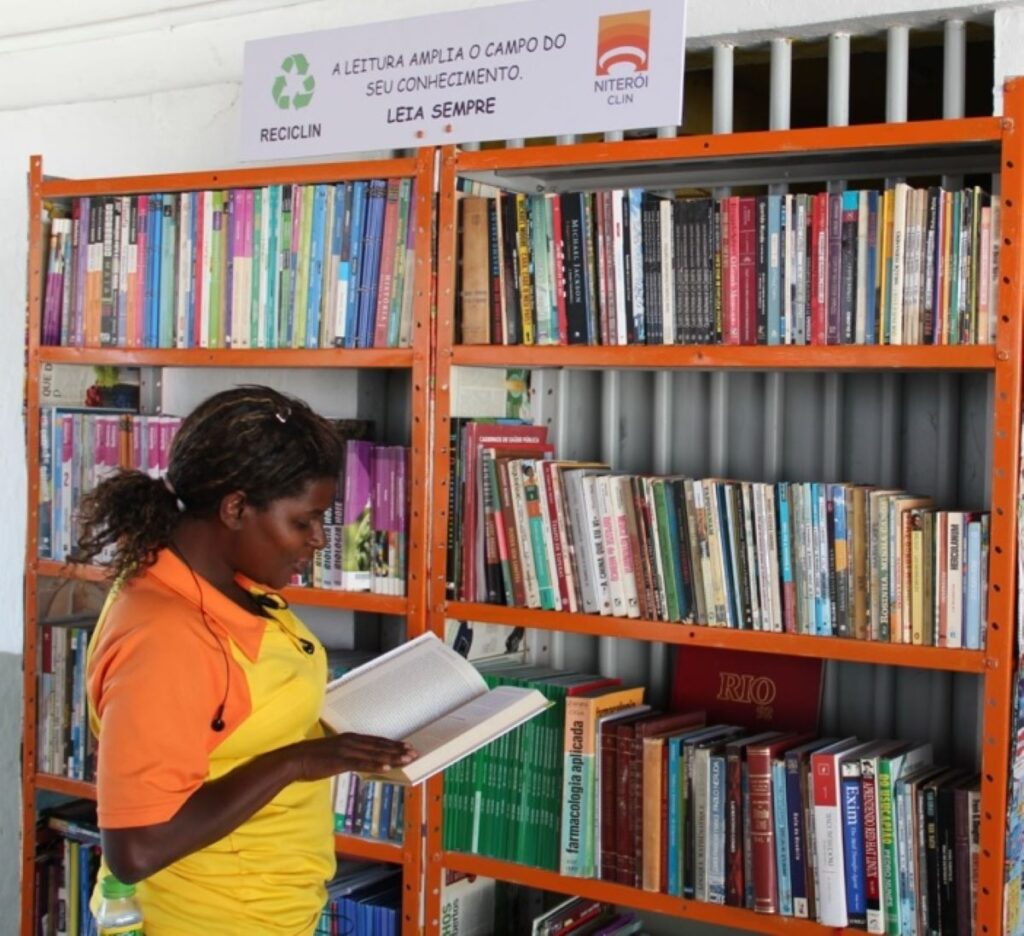Pessoas interagindo com estante de livros em biblioteca da CLIN em Niterói, parte do programa de circulação de obras.