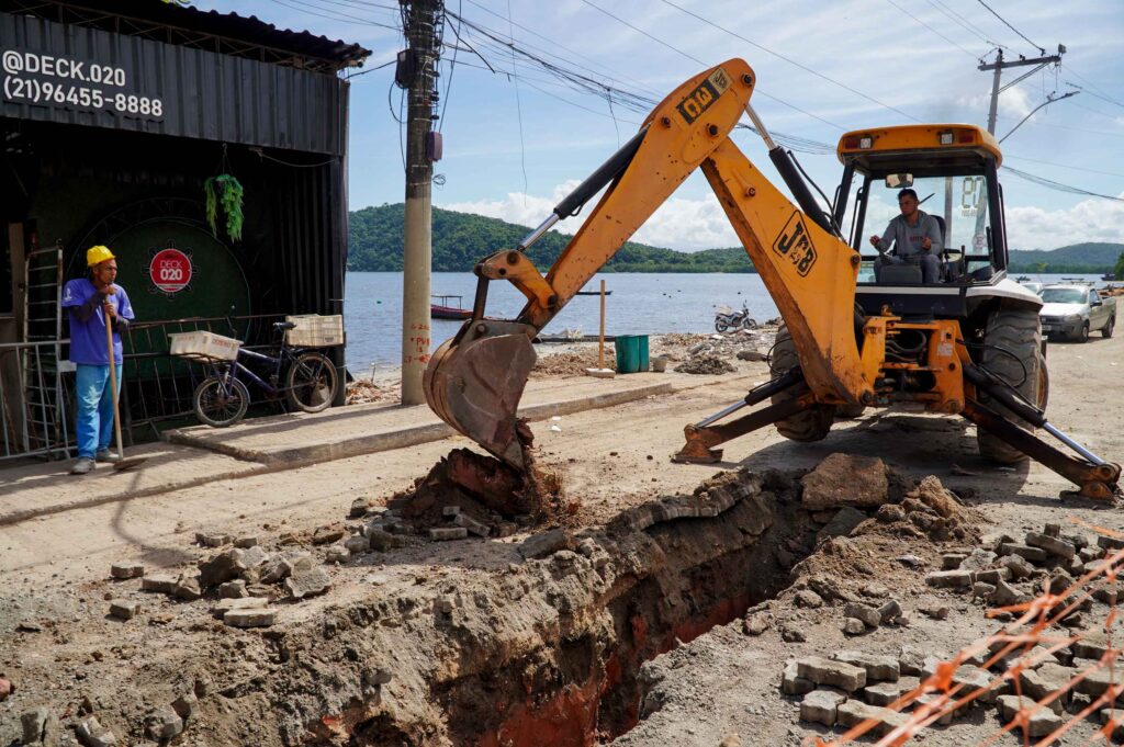 Obras de revitalização na orla da Praia das Pedrinhas, em São Gonçalo, com movimentação de máquinas e trabalhadores.