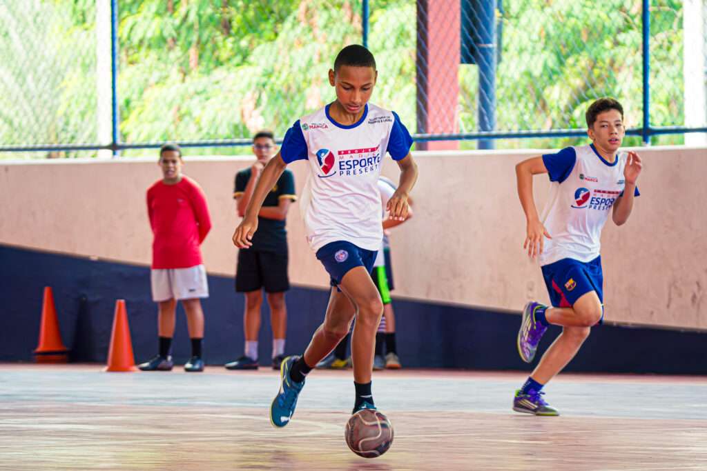 Adolescentes praticando futsal em quadra, com um professor orientando.