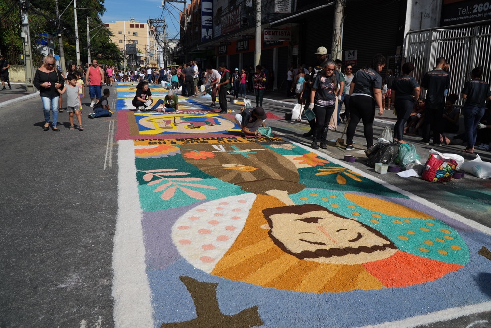 Tapetes de sal coloridos em rua de São Gonçalo durante celebração de Corpus Christi, com fiéis ao fundo.