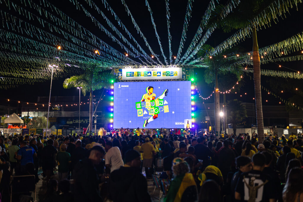Torcedores assistem a jogo da Copa do Mundo em telão ao ar livre em Maricá, com bandeiras do Brasil.