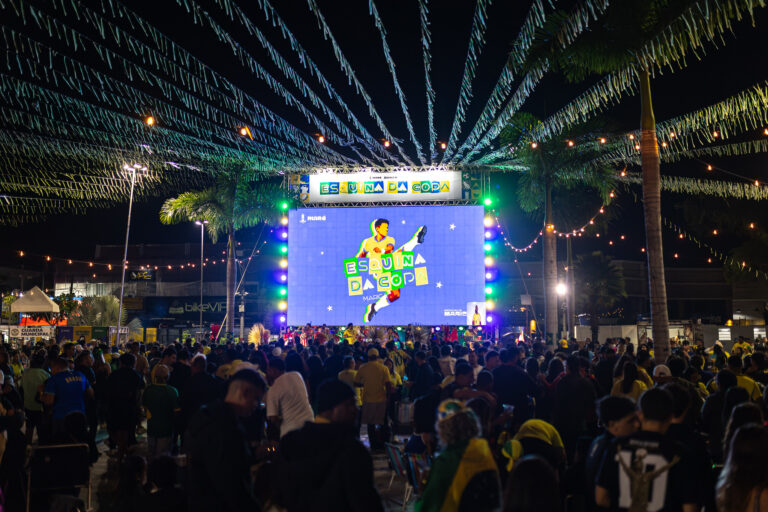 Torcedores assistem a jogo da Copa do Mundo em telão ao ar livre em Maricá, com bandeiras do Brasil.