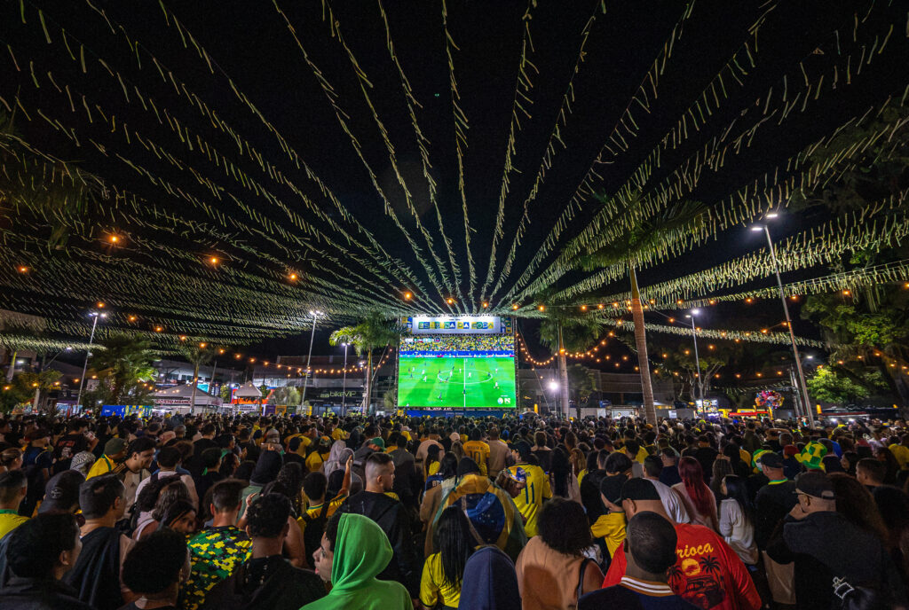 Pessoas reunidas em praça de Maricá assistem a jogo do Brasil em telão, com bandeiras e clima de festa.