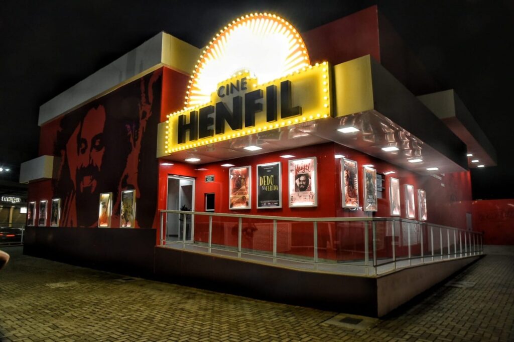 Pessoas assistem a um filme na sala escura do Cine Henfil em Maricá, com a tela iluminada mostrando uma cena.