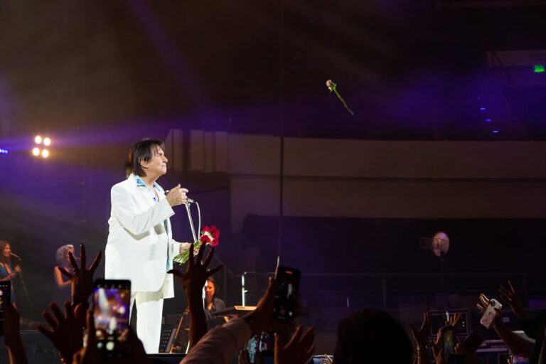 Roberto Carlos cantando em show, com microfone na mão e sorriso no rosto. Iluminação azul no fundo.