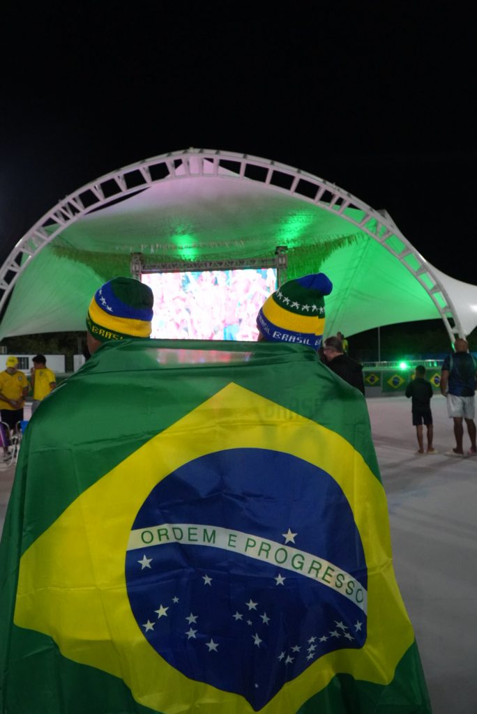 Telão transmitindo jogo de futebol no Parque RJ Nosso Sonho em São Gonçalo, com público ao redor e DJ.