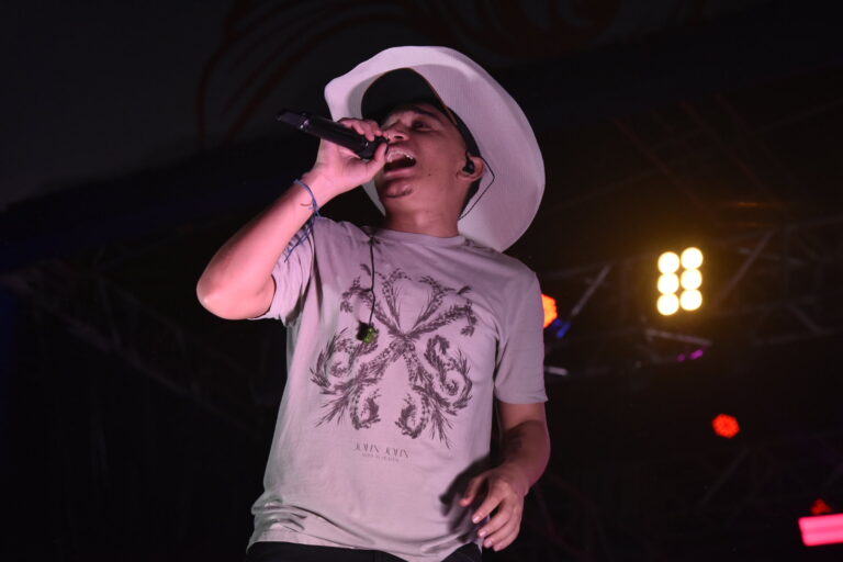 João Gomes cantando em show, com microfone na mão e palco iluminado.