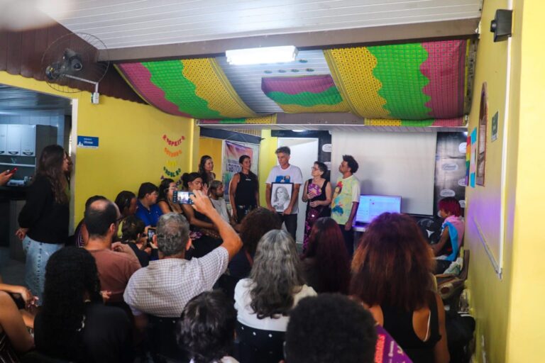 Mulher falando em microfone, com outras pessoas ao fundo, simbolizando cuidado vocal e comunicação.