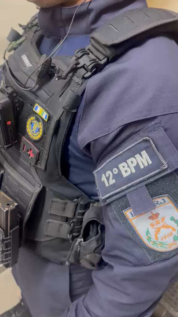 Policiais militares em operação em comunidade, com viatura ao fundo, representando ação policial.