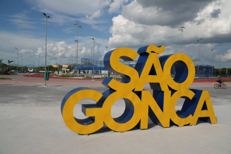 Pessoas jogando handebol de praia em quadra de areia do Parque RJ Nosso Sonho em São Gonçalo, com público ao redor.