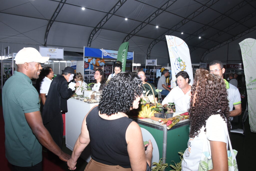 Pessoas em estandes de feira de negócios em Maricá, com logotipos de empresas e Prefeitura ao fundo.