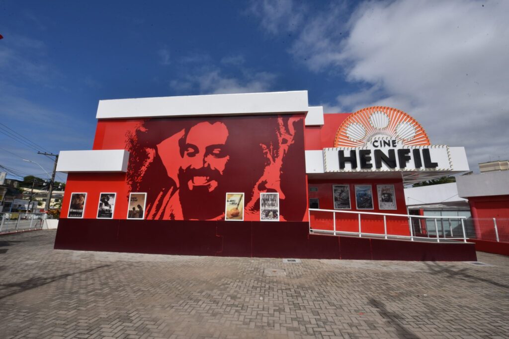 Público assiste a filme em sala escura do Cine Henfil em Maricá, com tela iluminada e poltronas vazias.