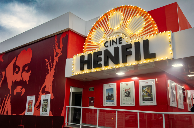 Pessoas assistindo a um filme em uma sala de cinema, com a tela iluminada e cadeiras vazias à frente.