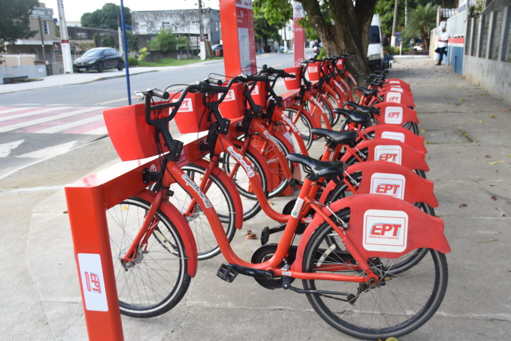 Pessoas utilizando bicicletas vermelhas do sistema público de Maricá em uma ciclovia na orla da cidade.