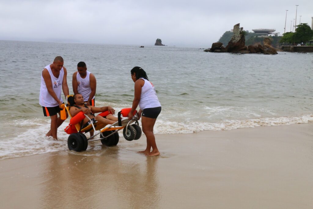 Pessoas com nanismo e cadeirantes participam do projeto Praia Sem Barreiras em Niterói, com acesso facilitado à areia e ao mar.
