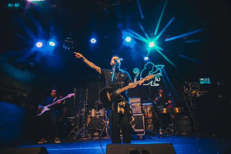 Cena de churrasco ao ar livre com público e palco de shows em Camboinhas, Niterói, ao pôr do sol.