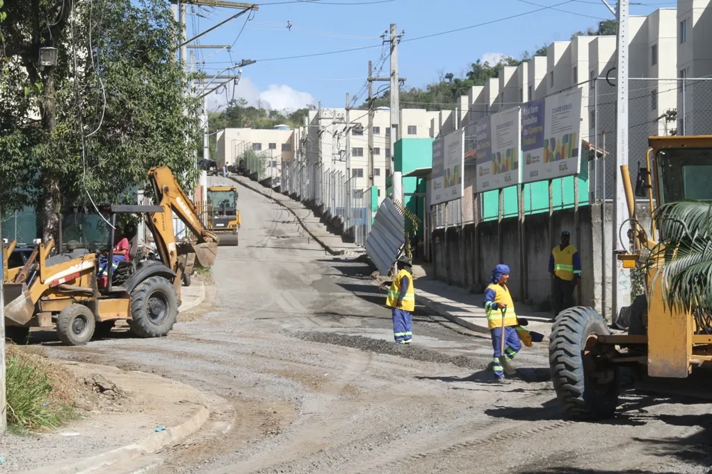 Obras de pavimentação e construção de calçadas em rua de São Gonçalo, com moradores observando o progresso.