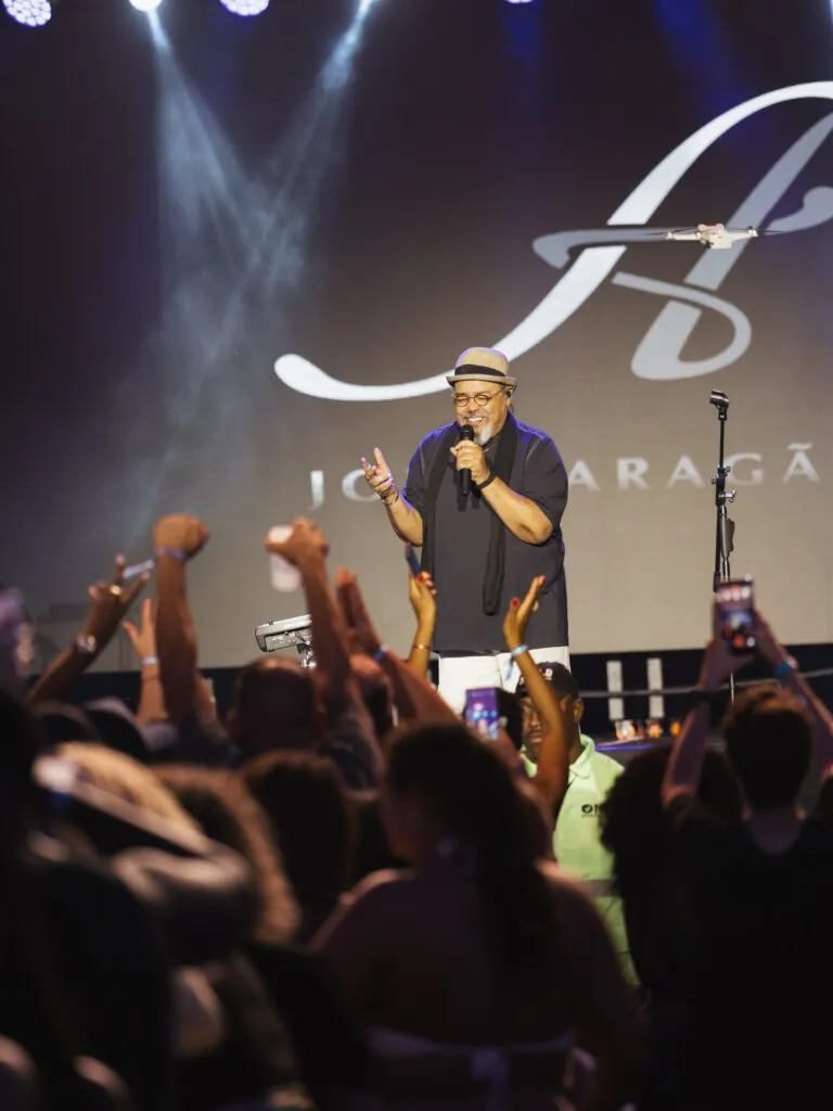 Jorge Aragão cantando em palco com microfone, iluminado por luzes coloridas, durante show da Festa do Trabalhador em Niterói.
