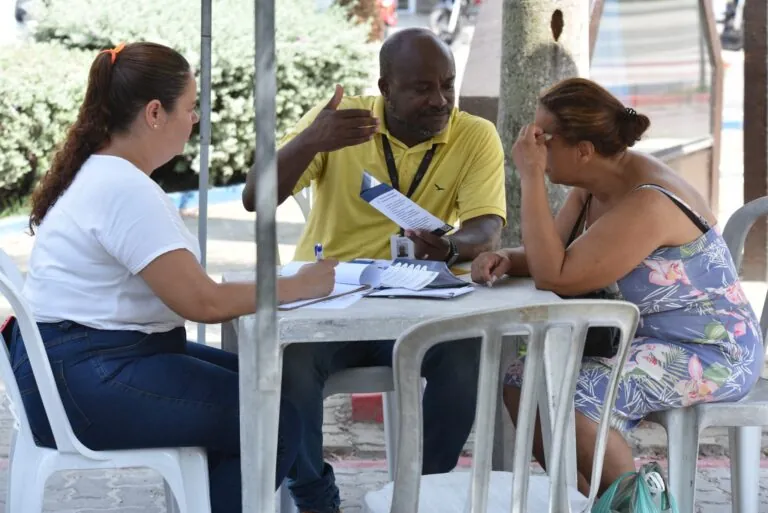 Pessoas sendo atendidas em balcão do Procon Itinerante em Maricá, com cartazes de orientação sobre direitos.